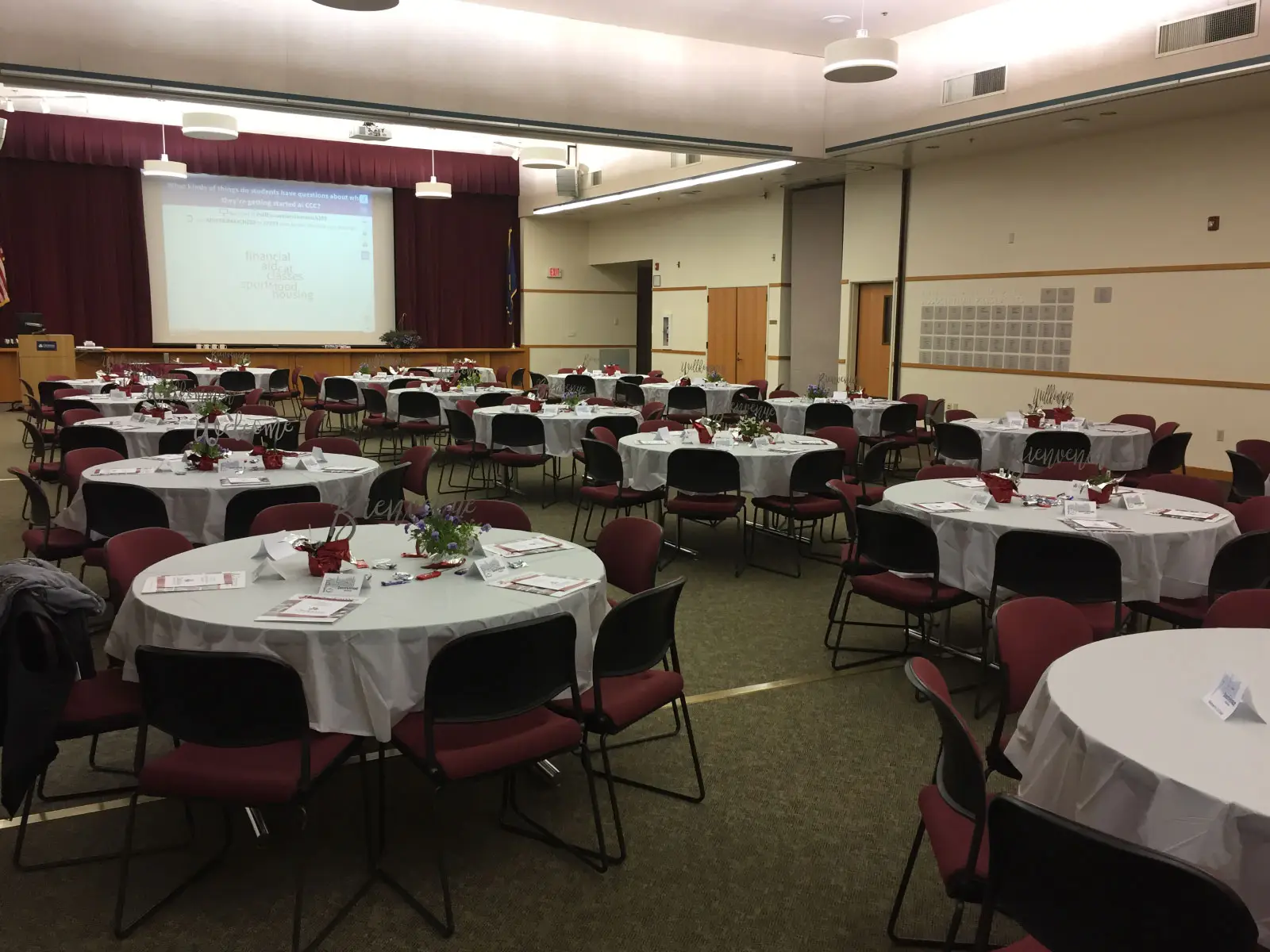 Many tables with red chairs, table centerpieces and silverware with a projector screen at the front of the room
